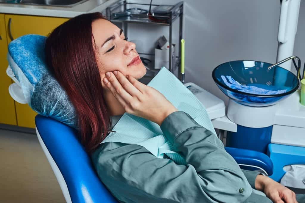 Woman sitting in dental chair holding her cheek with a pained expression